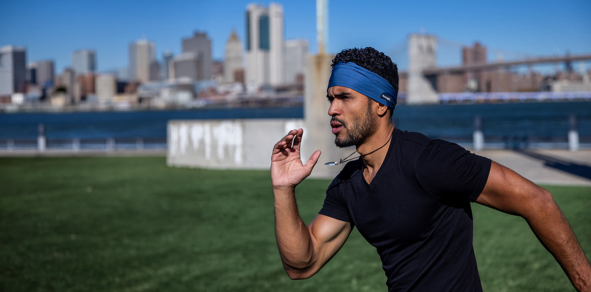 Man running outdoors in a blue Good Bands sweatband and black tee with the Brooklyn Bridge and Manhattan skyline behind him
