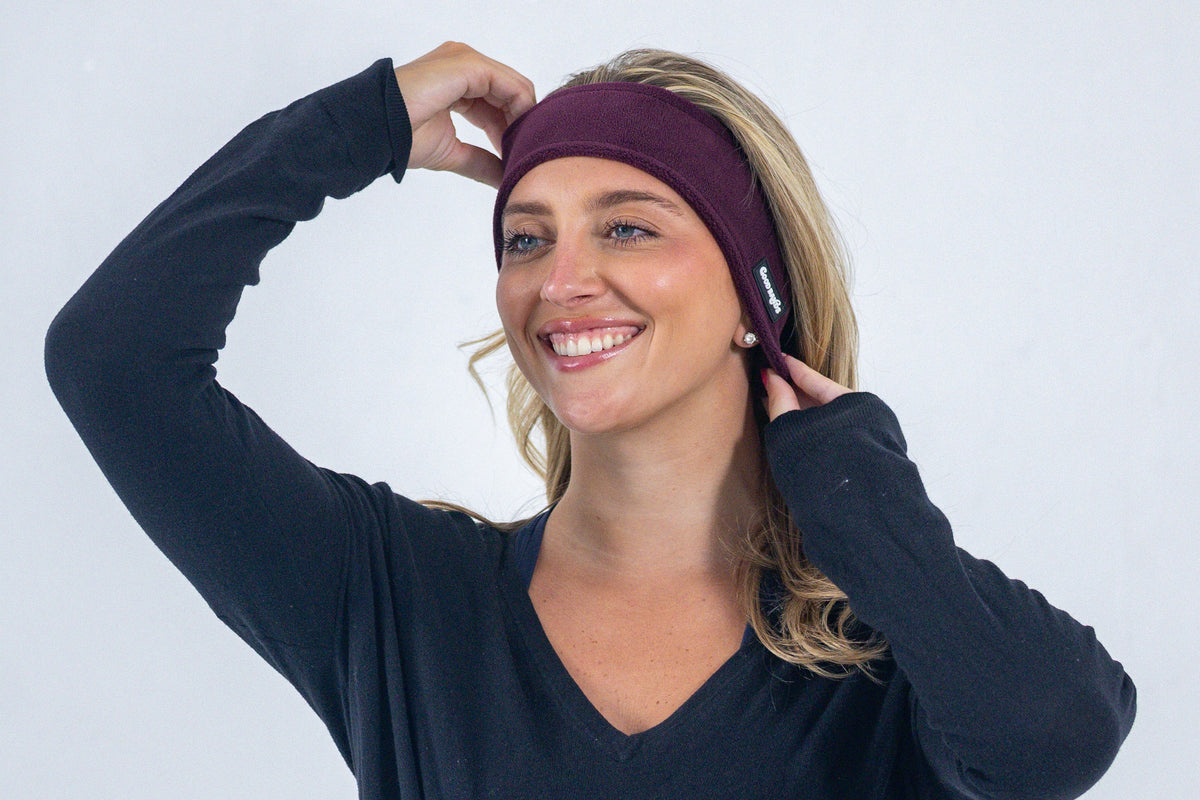 Smiling woman adjusting a plum purple Good Bands winter ear warmer, wearing a black long-sleeve V-neck on a light gray background