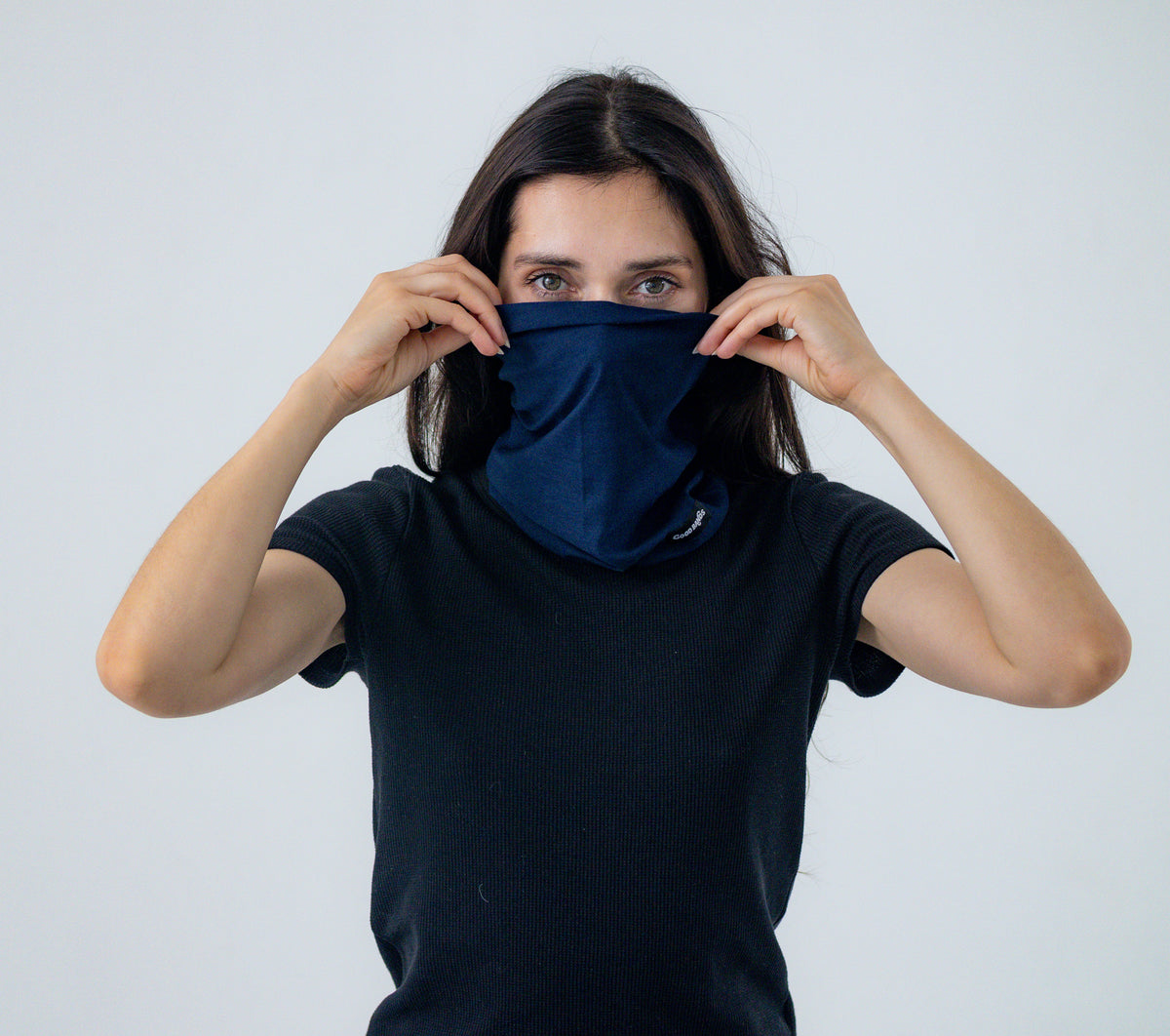 Woman pulling a navy blue Good Bands neck gaiter up over her nose and mouth as a face cover, on a light gray background
